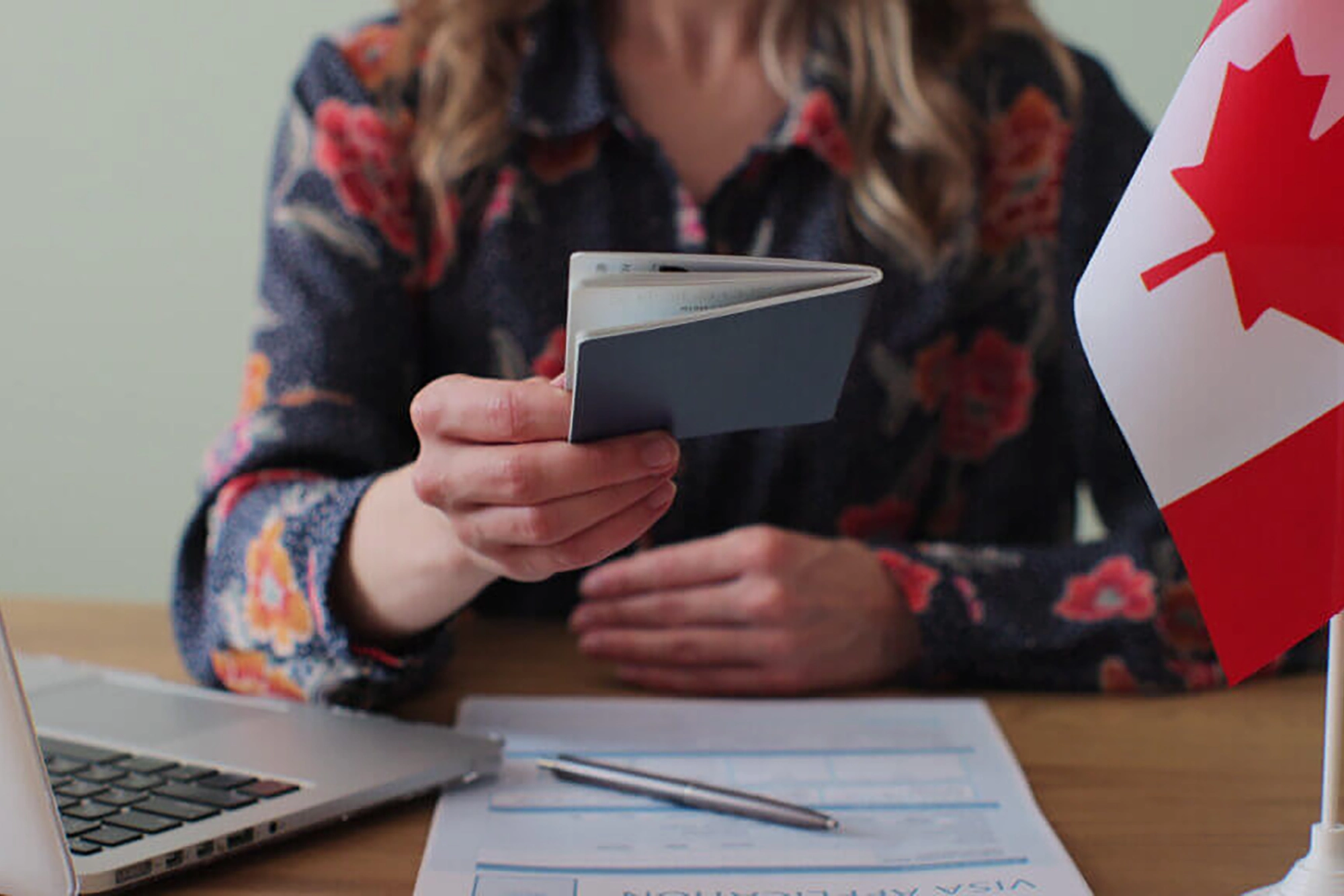 A woman across a desk is holding a Canadian passport, with the Canadian flag showing to the right of the image.