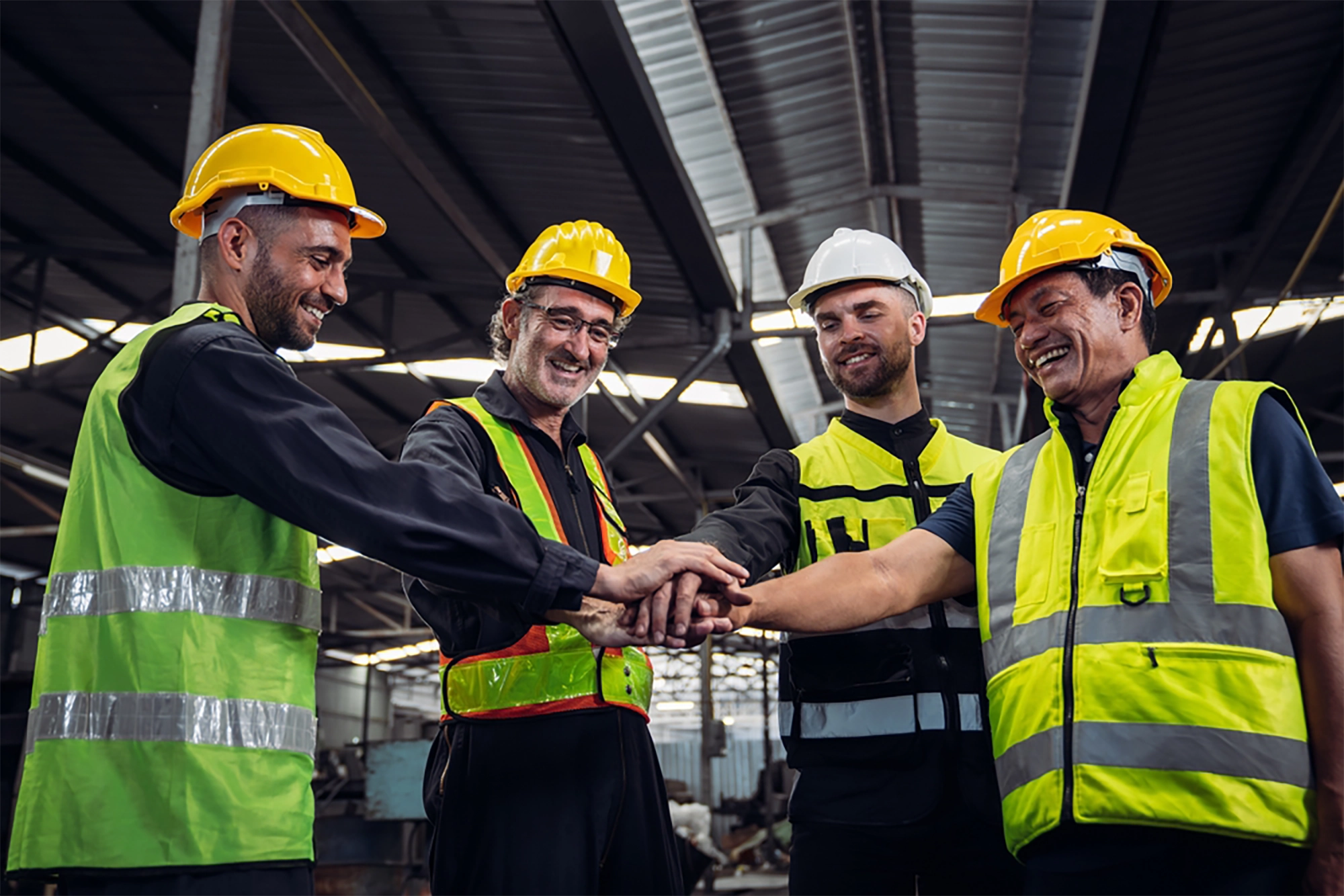 A group of four construction workers wearing safety helmets and reflective vests join hands in a gesture of teamwork inside a factory or warehouse setting.