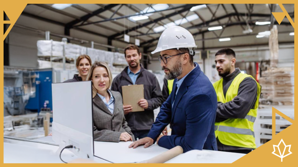 A group of workers and professionals in a manufacturing facility is discussing international recruitment, with a man in a hard hat and suit leading the conversation while looking at a monitor.