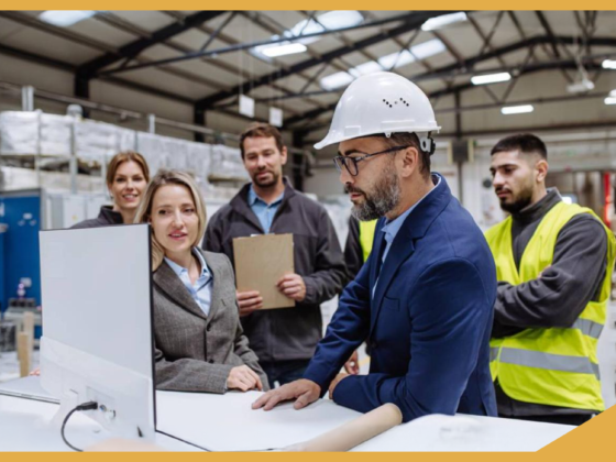 A group of workers and professionals in a manufacturing facility is discussing international recruitment, with a man in a hard hat and suit leading the conversation while looking at a monitor.