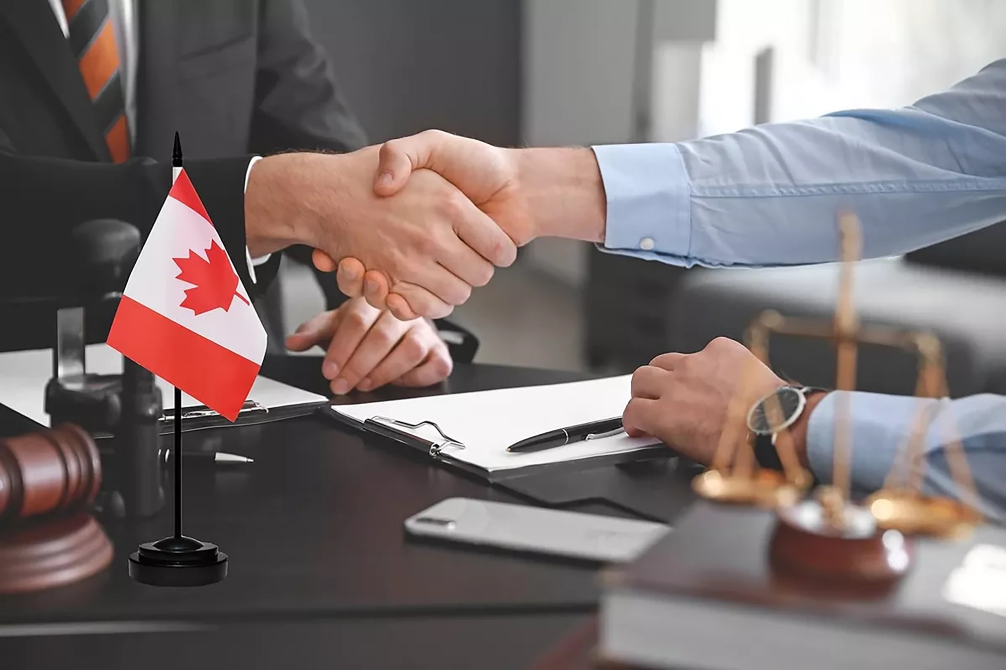 Two individuals in a formal office setting shaking hands across a desk with a small Canadian flag in the foreground, symbolizing an international agreement or partnership.