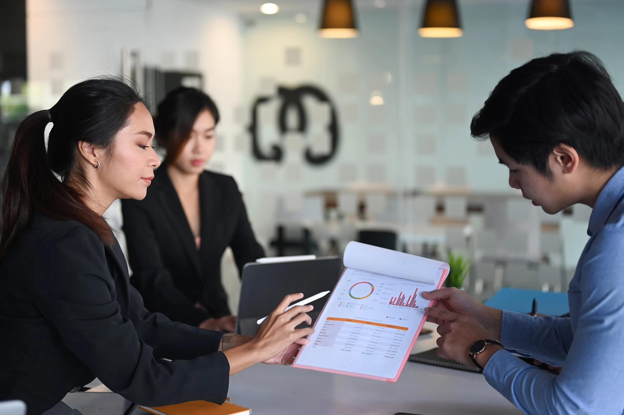 A group of three business professionals having a discussion about international recruitment in a modern office, with one person pointing to charts and graphs on a document.