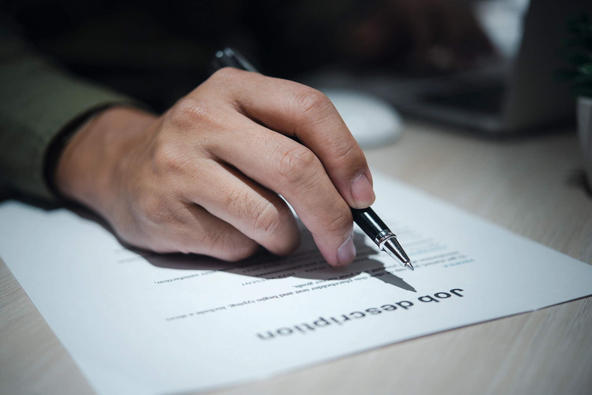 A close-up of a hand holding a black pen, writing or reviewing a job description document on a desk, with a laptop slightly blurred in the background.