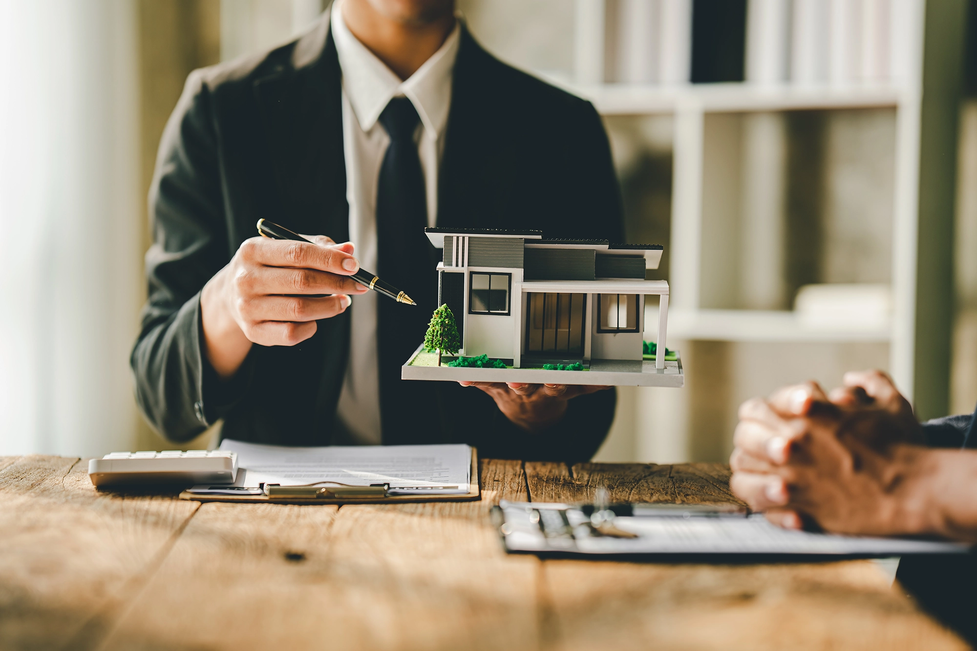 A real estate agent presenting a miniature model of a house to a client during a meeting, with documents and a calculator on the table.