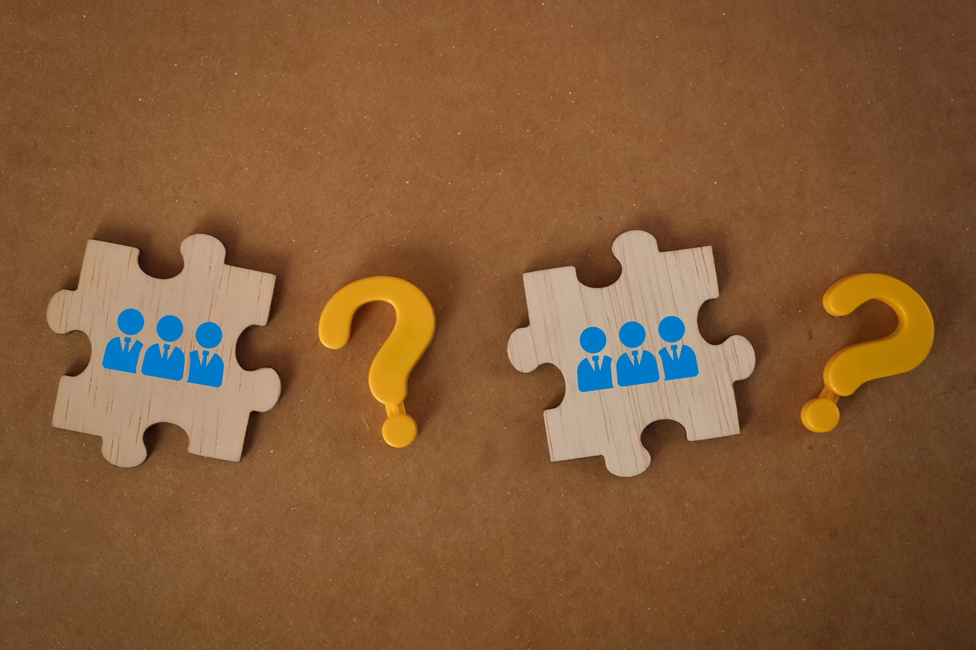 Two wooden puzzle pieces with blue icons of business teams, separated by yellow question marks, symbolizing the challenge of connecting and recruiting international teams.