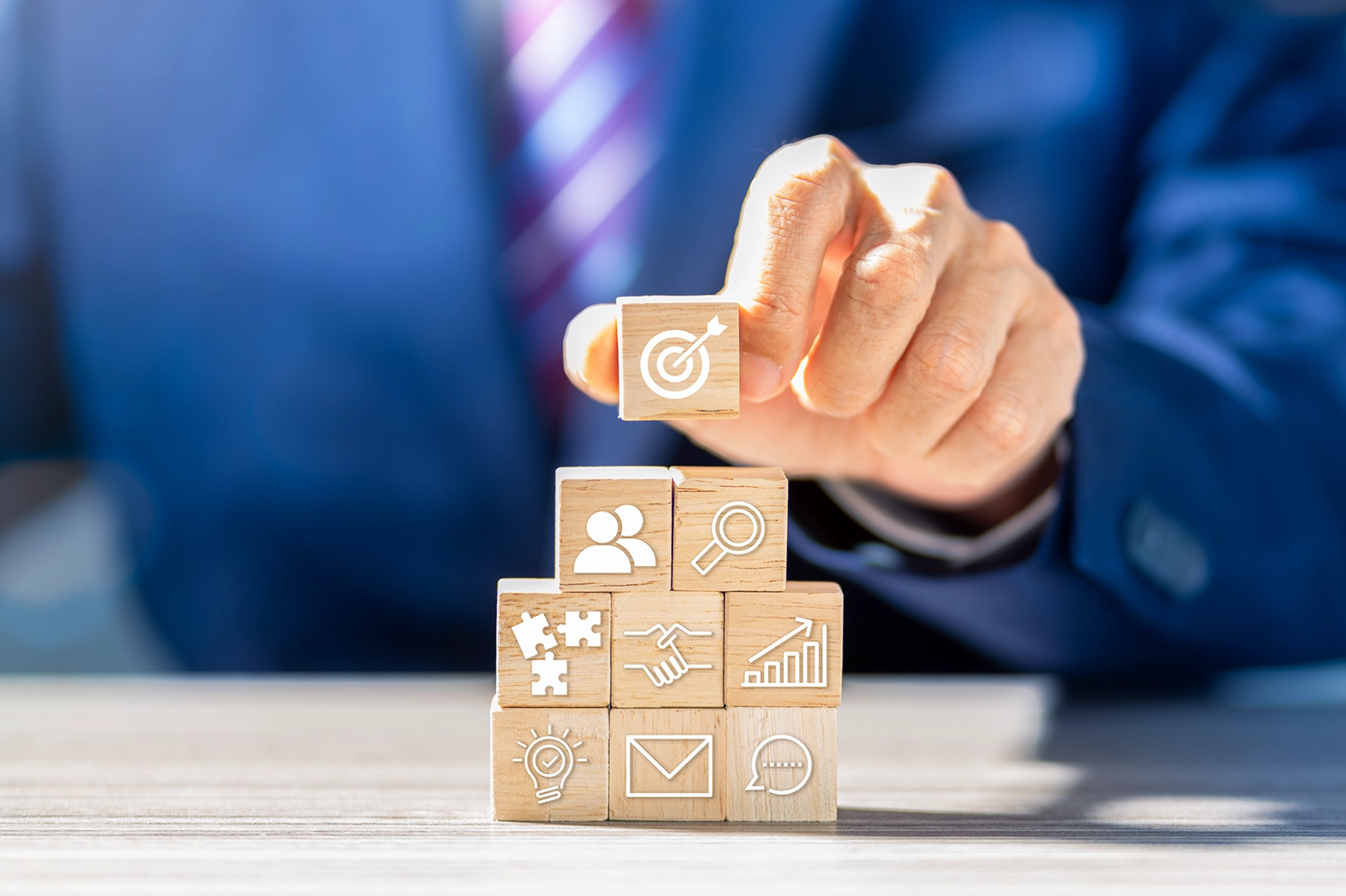 A person in a suit stacking wooden blocks with icons representing teamwork, goals, growth, and communication, illustrating strategic planning in international recruitment.